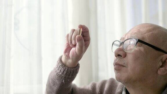 一位中国老人坐在窗边手里拿着一个胶囊戴着老花镜看着