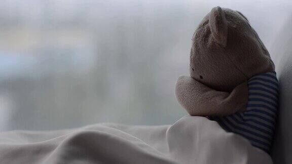 泰迪熊独自在床上哭泣他的白色枕头和毯子靠近窗户在早晨的悲伤的一天的雨滴
