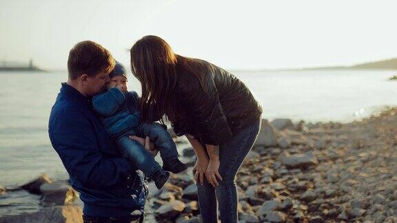 爸爸把小儿子抱在怀里和妻子一起亲吻他快乐国际一家人沿海岸散步肖像的观点