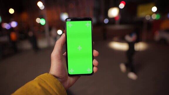 POV男性在夜晚的城市中手持绿色屏幕的智能手机