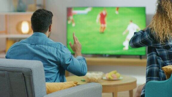 在家里一对年轻的夫妇在电视上看足球比赛当他们的球队进球时他们情绪上担心欢呼和庆祝胜利后视图相机拍摄