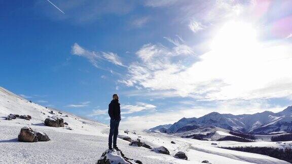 鸟瞰图年轻的白种人胡子时髦的男人站在一个巨大的石头上的风景如画的雪山覆盖的岩石晴天蓝天白云