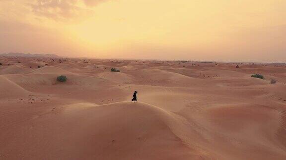 一架无人机在一名身穿阿拉伯联合酋长国传统服装的女子旁边的空中俯瞰她正在沙漠中的沙丘上行走