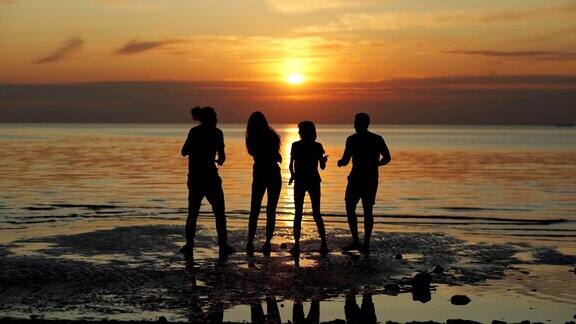 四个不同的年轻朋友的剪影与啤酒瓶在海上在橙色日落海滩派对背光的背影