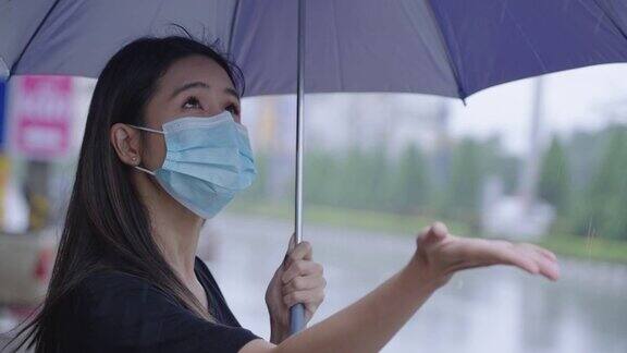 年轻的亚洲女子戴着外科口罩撑着雨伞站在路边在下雨的日子阵雨的热带雨季伸出手去感受雨滴保护自己
