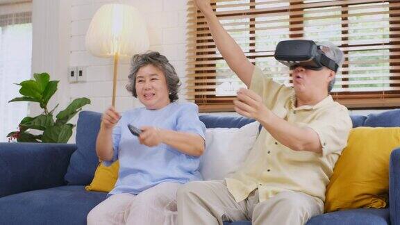 一对亚洲老年夫妇坐在家里的沙发上戴着虚拟现实眼镜拿着遥控器高级快乐时刻