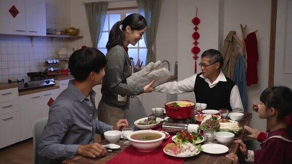 笑容可掬的亚洲儿媳拿着火锅把它放在便携式炉子上快乐的老爸爸打手势坐在餐桌前准备开始吃团圆饭