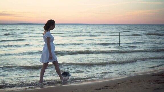 迷人的年轻女子在日落时沿着海滩散步一个穿着白色裙子的女孩赤脚沿着海滩走