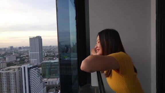 4K亚洲女人放松在公寓阳台看日落的城市风景