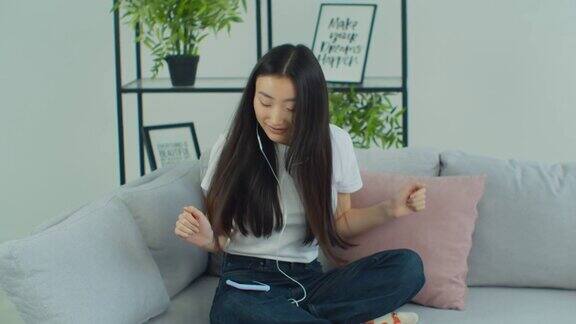 一个年轻的亚洲女孩坐在沙发上戴着耳机听音乐跳舞放松的年轻女子听最喜欢的音乐耳机在家里