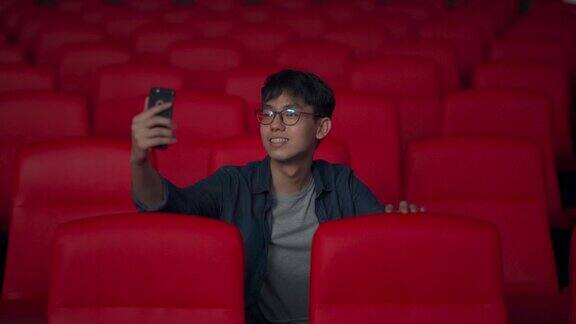 一名亚裔中国年轻人一边用智能手机自拍一边在电影院等待电影放映时间一边在社交媒体上发帖