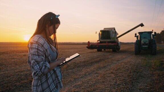 日落时分年轻的女农民一边看联合收割机将谷物卸到拖车上一边使用数码平板电脑