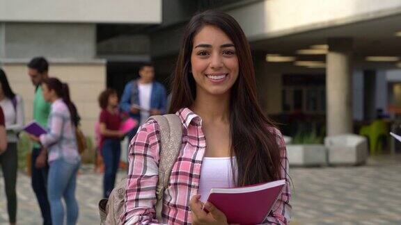 拉丁美洲美丽的学生拿着她的笔记本看着相机微笑着在大学校园