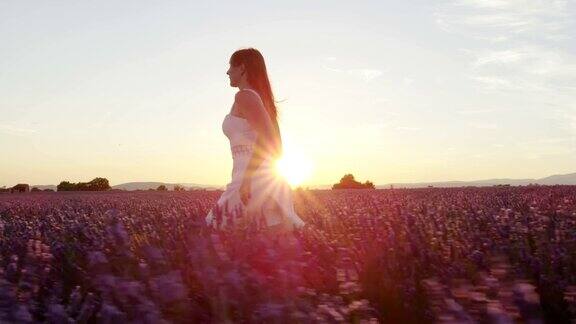 慢镜头:在金色的夏日落日中身着白裙的快乐年轻女子奔跑在美丽的紫色薰衣草田中
