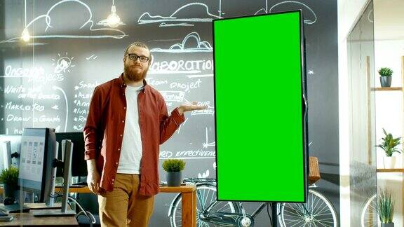 男性开发者看着带有绿色屏幕模型的交互式白板他微笑着用手势指着屏幕在背景创意办公室与黑板墙与项目计划绘制