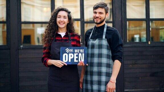 美丽的女士和她的商业伙伴英俊的男人举着开放的标志通知顾客开放他们的咖啡馆和欢迎的人的慢动作肖像