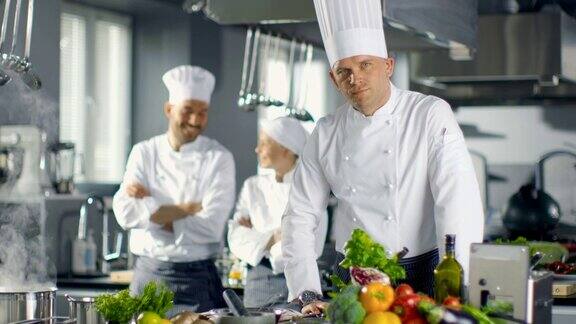 一家大餐厅的名厨准备菜肴在镜头前微笑背景中的两个学徒和现代厨房