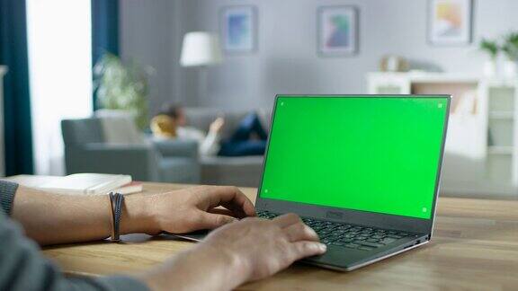 男人的手在绿色模拟屏幕的笔记本电脑上打字在舒适的客厅里一个女人在沙发上休息