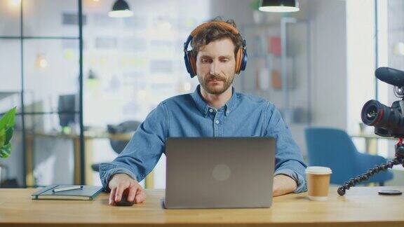 年轻英俊的男人在一个阁楼办公室的酷创意机构的笔记本电脑上工作他有一杯外卖咖啡桌上有一本笔记本他穿着牛仔裤衬衫戴着耳机