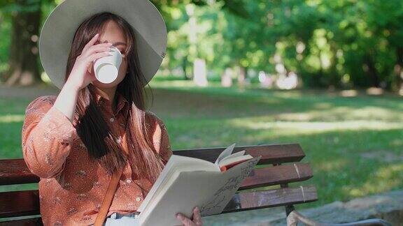 一个年轻的女人正坐在公园的长椅上一边看书一边喝咖啡
