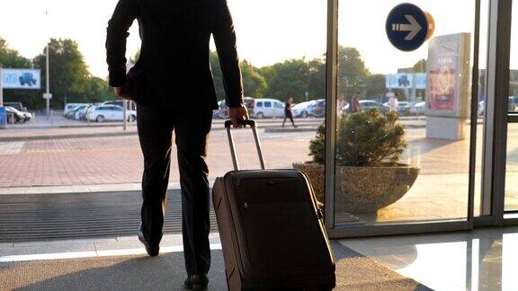 一名年轻人从机场的玻璃自动门进入市区拉着带轮子的行李箱商人看了看他的手表带着行李从航站楼走向停车场后视图慢动作
