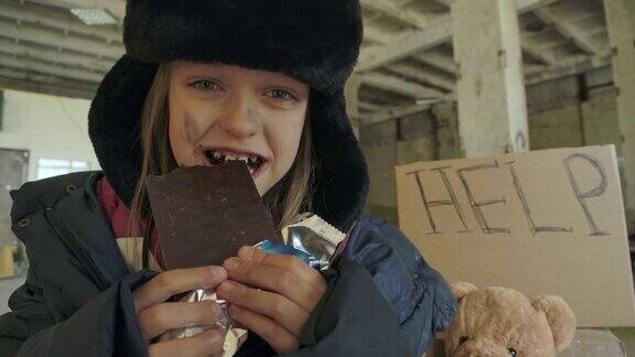 微笑的白人无家可归的女孩肮脏的脸和灰色的眼睛贪婪地吃巧克力棒