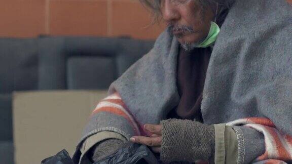 4K戴着医用口罩的无家可归老人坐在城市的人行道上