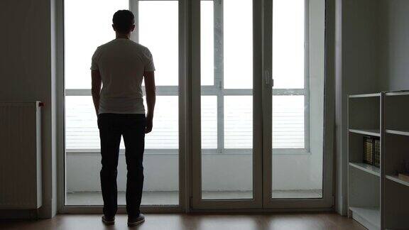 一个悲伤的年轻人的剪影站在家里的大窗户旁边