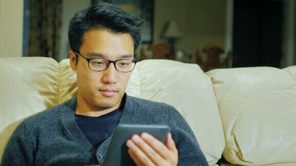 一个戴眼镜的亚洲年轻人他坐在家里的沙发上看电子书