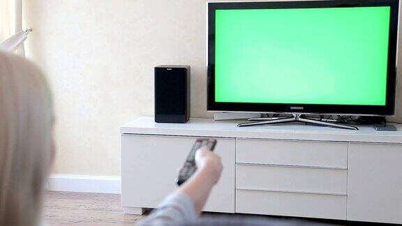 一个金发女郎坐在客厅的沙发上用电视控制器看绿色屏幕的电视