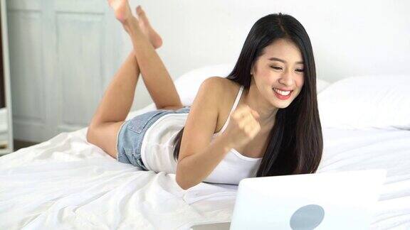 美丽的亚洲年轻女子躺在床上使用笔记本电脑在卧室的休闲和放松与成功自由职业者与女孩工作笔记本与兴奋沟通慢动作