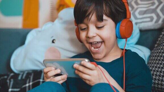 小男孩玩手机游戏愉快地玩手机游戏他使用耳机以避免受到干扰5岁男孩在玩手机游戏学龄前儿童在小工具上玩游戏