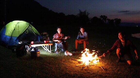 一群亚洲朋友游客正在玩尤克里里而他的放松的朋友正在听和扔柴火篝火坐在一起附近的帐篷