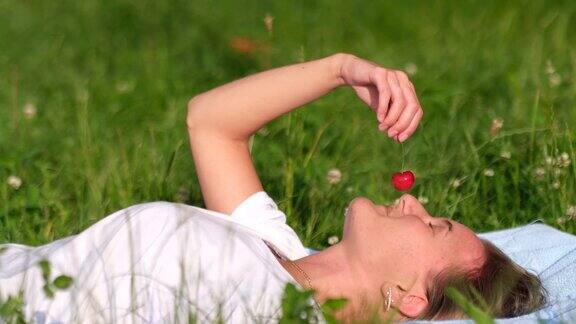 一个年轻的女孩躺在绿色的草地上品尝着一颗成熟的红樱桃