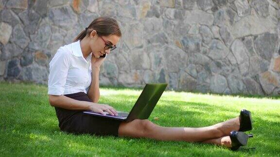 女商人坐在公园的草地上拿着笔记本电脑接电话