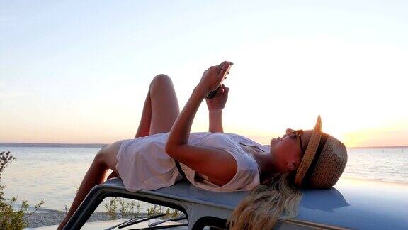 女孩在车顶上自拍日落旅行手机年轻女子躺在车顶上自拍手机