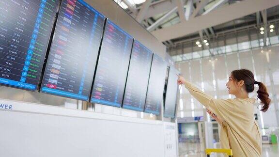 年轻的亚洲女子拿着护照和登机牌在国际机场看航班信息板检查她的航班