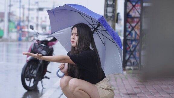 一个漂亮的亚洲女孩撑着伞坐在路边散步在下雨的日子里她伸出手触摸着雨滴弄坏了摩托车失望地困在雨中