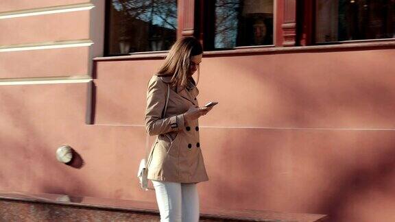 身材苗条的年轻女子穿着米色夹克和牛仔裤在城市里走来走去看着手机慢动作