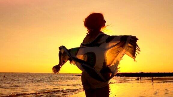 慢镜头:日落时分戴着太阳镜和飘动的围巾的女孩在海滩上旋转