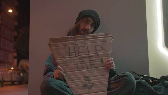 一个留着胡子和长发的无家可归的乞丐坐在街上寻求帮助的慢动作拿着“帮我”的牌子无家可归4k