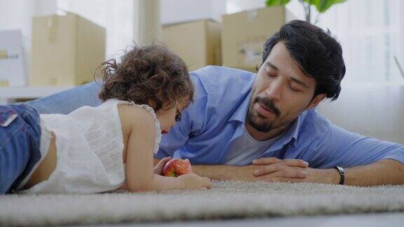 父亲和一头卷发的快乐小女婴躺在地板上在女儿吃东西的时候父亲一边说着话一边轻轻地抚摸着女儿的头一边咬着苹果准备搬进新家背景是纸箱