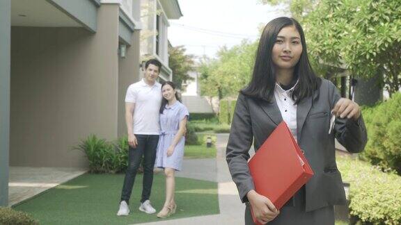 亚洲自信的女人房地产经纪人或房地产经纪人在西装拿红色文件和显示房子的钥匙与年轻夫妇的房屋卖家在房子前面一个可爱的女经纪人的肖像