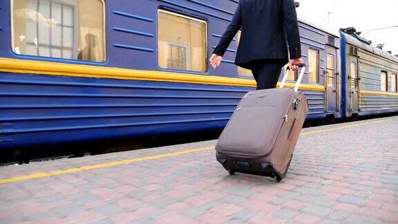 穿着黑色正装的成功商人沿着站台走着用轮子拉着手提箱自信的年轻人带着行李在路过的火车旁漫步慢镜头