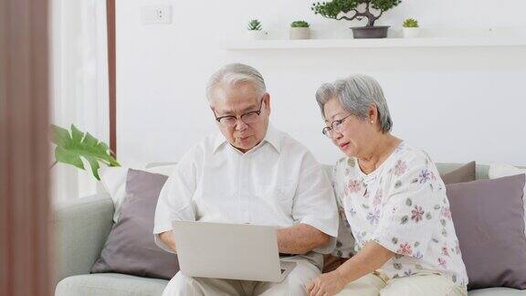 亚洲老年夫妇在家中客厅使用电脑笔记本上网搜索、网上购物、购买保险老年人具有科技理念
