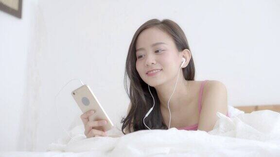美丽的亚洲女人躺在幸福的耳机听音乐与放松和享受在卧室女孩好玩的耳机玩智能手机生活理念慢动作