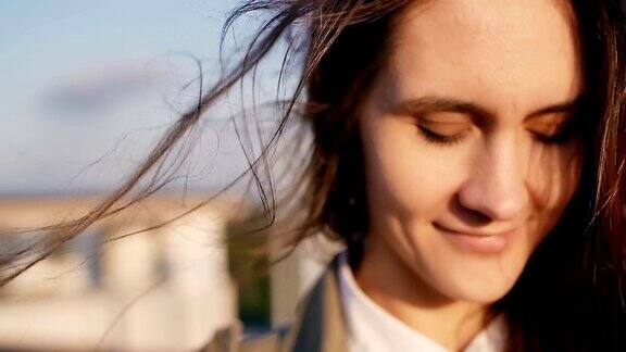 风神奇的吹着长发的女孩幸福微笑的女人站在屋顶上看夕阳看着相机缓慢的莫