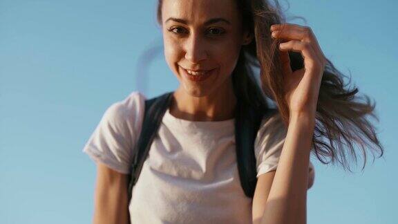 身穿白色t恤的年轻漂亮快乐的女子站在蓝天的背景上用手拉直头发看着镜头平静和放松