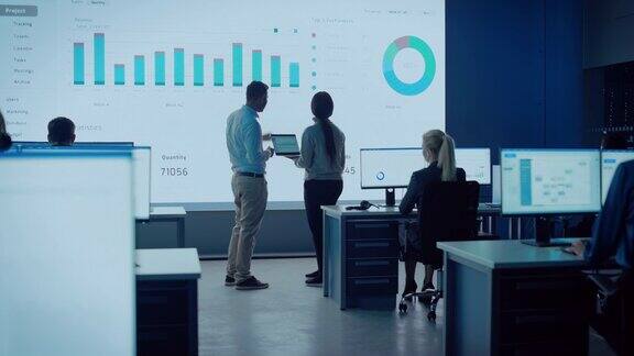 两个交易员正在一个现代的监控办公室里开会大屏幕上显示着分析数据有经纪人和金融专家坐在电脑前的监控室同事们说话