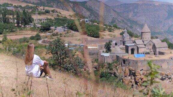 在亚美尼亚一个女孩正坐在群山的背景下塔捷夫修道院的教堂建筑风吹着头发背景是中世纪的城堡